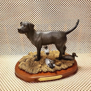 May include: A dark brown Labrador retriever statue stands on a base with a duck decoy and two small ducks. The base is oval-shaped and made of wood, with a gold-colored plaque that reads "THIRD SEASON".