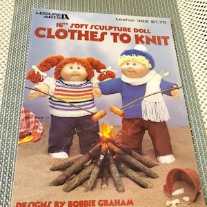 May include: A knitting pattern booklet for a 16-inch soft sculpture doll. The cover features two dolls wearing knitted clothes, sitting by a campfire. The title of the booklet is "Clothes to Knit" and the text "Designs by Bobbie Graham" is at the bottom.