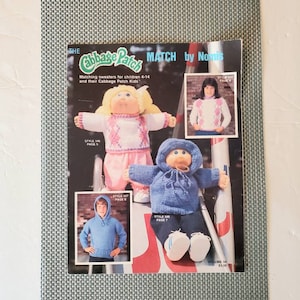 May include: A vintage Cabbage Patch Kids pattern book titled "MATCH by Nomis." The cover features images of Cabbage Patch Kids dolls and children wearing matching sweaters. The book includes the text "Matching sweaters for children 4-14 and their Cabbage Patch Kids."
