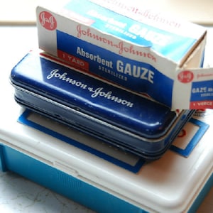 May include: A blue metal first aid kit with a white lid. The kit is labeled "Johnson & Johnson" and contains a roll of absorbent gauze labeled "Johnson & Johnson Absorbent Gauze Sterilized 1 Yard".