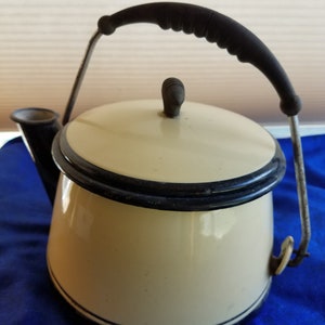 May include: A vintage white enamel teapot with a black trim and a black handle. The teapot has a spout and a lid.