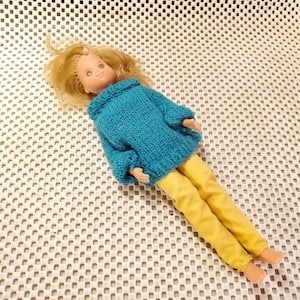 May include: A vintage doll with blonde hair, blue eyes, and a teal blue knitted sweater. The doll is wearing yellow pants. The doll is lying on a white textured surface.