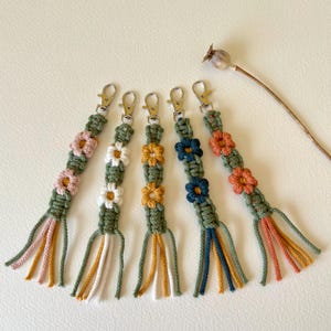 May include: Five macrame keychains with floral patterns. Each keychain has a different colour scheme, including pink, white, yellow, blue, and orange flowers. The keychains have a green base and matching tassels, and are attached to silver clasps.