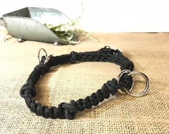 May include: A black paracord dog collar featuring two large silver metal rings and a woven pattern. The collar is knotted and appears robust. The background includes a hessian-style surface and a decorative metal container.