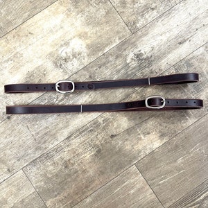 May include: Two brown leather straps with silver buckles. The straps are lying flat on a wood grain surface.