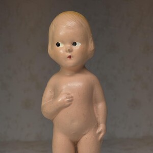 May include: A vintage bisque doll with a peach skin tone. The doll has a simple hairstyle, black eyes, and a red mouth. It is wearing no clothing and has molded socks. The doll is standing upright.