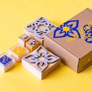 May include: A tile stamp set with various gray floral stamps on wooden blocks, alongside ink pads in yellow and blue. The set includes a cardboard box with a blue and yellow floral design and the text "TILES STAMP SET".