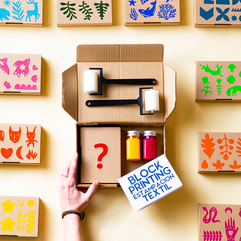 May include: A cardboard box filled with supplies for block printing. The box contains two rollers, two bottles of paint, and a booklet with the text "Block Printing Estampacion Textil". The box is surrounded by small cardboard boxes with various colourful designs.