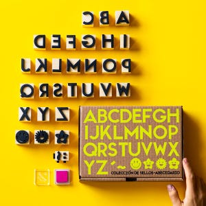 May include: A set of alphabet stamps with wooden blocks and black letters, arranged on a bright yellow background. The set includes a cardboard box with the alphabet in green, along with ink pads and decorative stamps. The text on the box reads "COLECCIÓN DE SELLOS-ABECEDARIO".