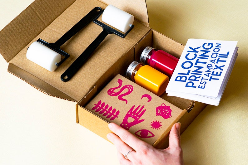 May include: An open cardboard box containing a block printing kit. The kit includes two bottles of fabric paint, one yellow and one pink, two white rollers, a black roller handle, and a sheet of pink fabric stamps with designs such as a snake, a hand, a crescent moon, and a sun.