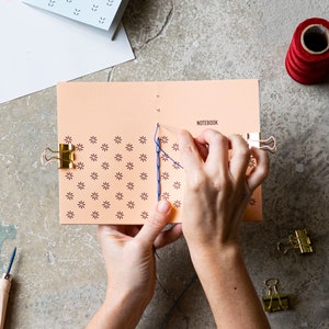 Pode incluir: Um caderno cor de pêssego com um padrão floral está sendo costurado à mão com linha azul. A palavra "NOTEBOOK" está impressa no lado direito. Clipes dourados prendem as páginas no lugar. Um carretel de linha vermelha e outros artigos de papelaria estão por perto.