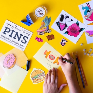 Könnte beinhalten: Ein Bastelset mit den Worten "Kit Mágico DIY PINS" und "Ilustra tus propios pins" auf gelbem Hintergrund. Das Set enthält eine Vielzahl von bunten Pins, Markern und einer transparenten Acetatfolie.
