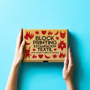 Op de afbeelding: Een bruine kartonnen doos met de tekst "BLOCK PRINTING ESTAMPACIÓN TEXTIL AITOR SARAIBA Y FABRICA DE TEXTURAS" erop gedrukt. De doos is versierd met rode harten, sterren en andere grafische elementen.