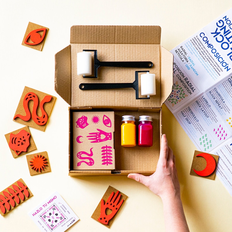 May include: An open cardboard box filled with art supplies for printmaking. The box contains two black and white ink rollers, two small bottles of paint, one yellow and one pink, and a sheet of pink and white stamps with various designs. The box is surrounded by a variety of colourful stamps, including a red eye, a red snake, a red hand, a red sun, and a red crescent moon.