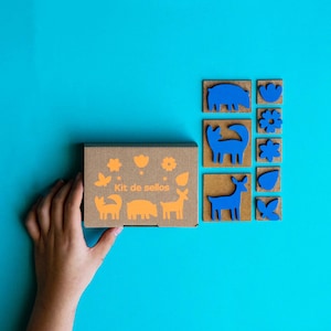 May include: A cardboard box with the text "Kit de sellos" on the front. The box contains a set of blue rubber stamps with various animal and flower designs.