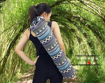 May include: A woman wearing black trousers carries a blue, brown and white patterned yoga mat bag with a drawstring closure. The bag is slung over her shoulder. The background is a lush green bamboo forest.