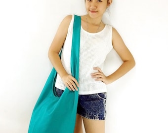 May include: A teal blue canvas tote bag with a long strap. The bag is worn over the shoulder of a person wearing a white tank top and denim shorts.