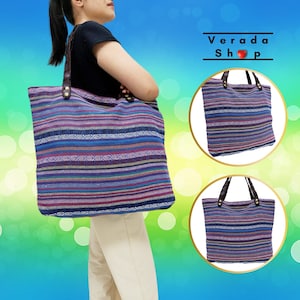 May include: A large, striped tote bag with brown leather handles. The bag is made of a woven fabric with a colourful pattern of blue, purple, and pink stripes. The bag is shown in two different views, one with a person holding it and the other with the bag laying flat.