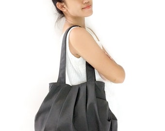 Puede incluir: Un bolso tote gris plisado con una sola correa. El bolso lo lleva una persona que lleva un top blanco sin mangas y pantalones cortos de mezclilla.