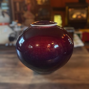 May include: A round, burgundy-colored ceramic urn with a glossy finish. The urn has a matching lid with a white rim. The piece sits on a wooden surface, showcasing its spherical shape and reflective surface.