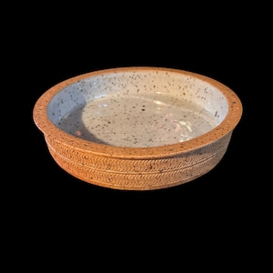 May include: A round, ceramic baking dish with a speckled, light gray interior and a textured, brown exterior. The rim features a repeating, wave-like pattern. The dish is designed for baking and serving.