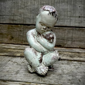 Puede incluir: Una escultura de cerámica de un niño sentado con la cabeza gacha, la escultura está hecha en un esmalte craquelado de color azul claro y blanco.