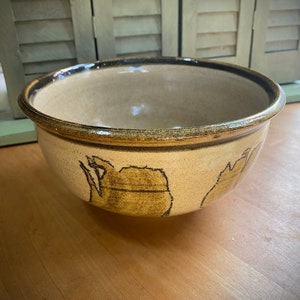 Puede incluir: Un tazón grande de cerámica marrón y crema con un borde marrón oscuro. El tazón tiene un diseño único de líneas y formas marrones sobre un fondo crema.