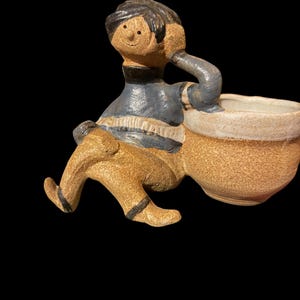 Könnte beinhalten: Ein skurriler Keramik-Pflanztopf in Form einer sitzenden Figur mit einem lächelnden Gesicht und dunklem Haar. Die Figur trägt ein blau-weiß gestreiftes Hemd und eine beige Hose. Der Pflanztopf ist beige mit einem weißen Rand.