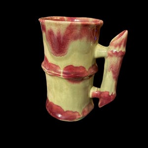 May include: A handmade ceramic mug with a bamboo-inspired design. The mug features a light green base with red accents and a matching handle. The mug is approximately 4 inches tall.