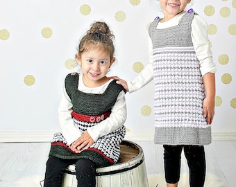 May include: Two young girls wearing crocheted dresses. The girl on the left is wearing a green and red dress with a white and black pattern. The girl on the right is wearing a gray and white dress with a purple stripe. Both girls are wearing black leggings.