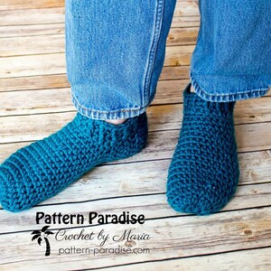 May include: A pair of teal blue crocheted slipper socks. The socks are worn by a person wearing blue jeans. The image includes the text "Pattern Paradise Crochet by Maria pattern-paradise.com".