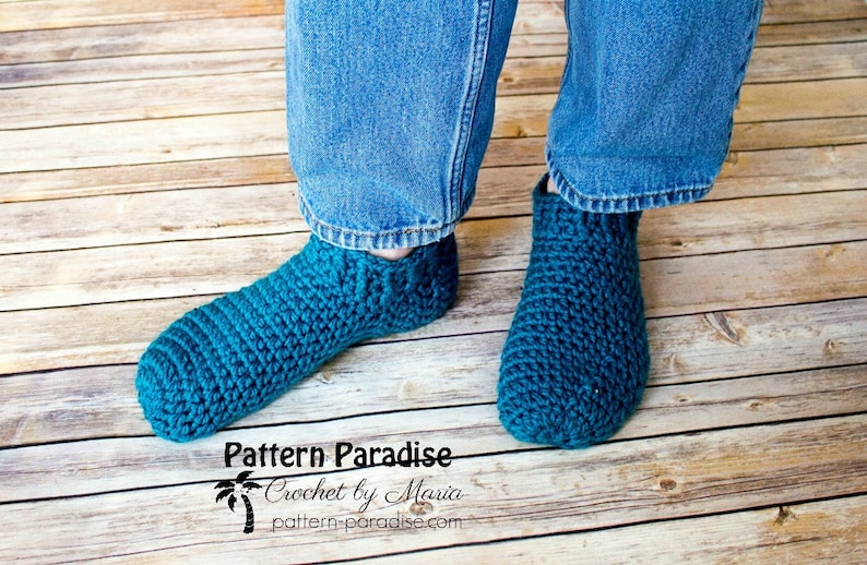 May include: A pair of teal blue crocheted slipper socks. The socks are worn by a person wearing blue jeans. The image includes the text "Pattern Paradise Crochet by Maria pattern-paradise.com".