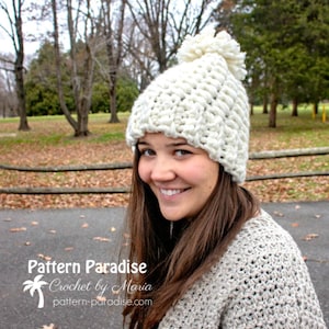 May include: A white crocheted beanie with a fluffy pom pom on top. The beanie is worn by a person with brown hair and a white sweater. The text "Pattern Paradise Crochet by Maria pattern-paradise.com" is visible at the bottom of the image.