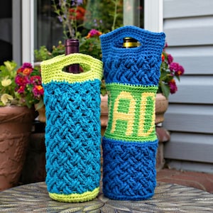 May include: Two crocheted wine bottle holders, one blue and green with a braided pattern, the other blue and green with a braided pattern and the word "YOU" in yellow yarn. The holders are sitting on a round, black, textured table.