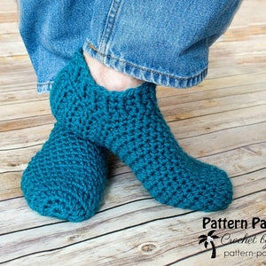 May include: A pair of teal blue crocheted slipper socks. The socks are worn on a person's feet. The socks are made with a simple stitch pattern.