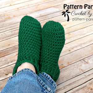 May include: A pair of green crocheted slippers worn on a person's feet. The slippers are made with a simple stitch pattern and have a ribbed cuff. The person is wearing blue jeans.