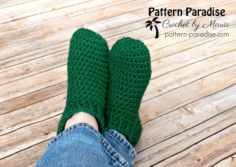 May include: A pair of green crocheted slippers worn on a person's feet. The slippers are made with a simple stitch pattern and have a ribbed cuff. The person is wearing blue jeans.