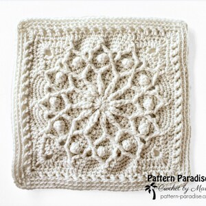 May include: Cream-colored, square crochet doily with a detailed floral design in the center. The doily features raised bobbles and a textured border. The text "Pattern Paradise Crochet by Maria" is visible in the lower right corner.
