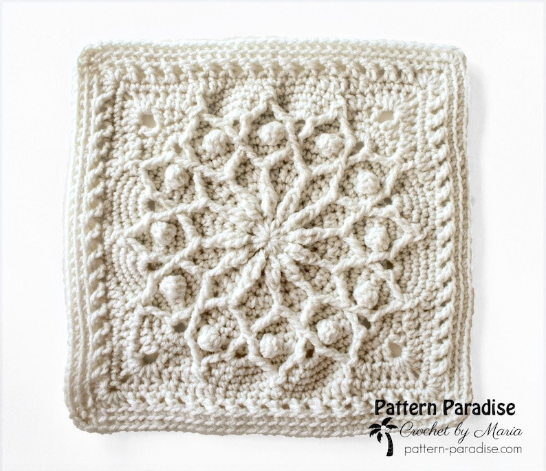 May include: Cream-colored, square crochet doily with a detailed floral design in the center. The doily features raised bobbles and a textured border. The text "Pattern Paradise Crochet by Maria" is visible in the lower right corner.