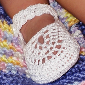 Könnte beinhalten: Weiße gehäkelte Babyschuhe mit einem zarten Spitzenmuster. Die Schuhe haben einen Riemen mit einem weißen Knopf.