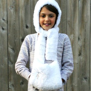 May include: A white crocheted hooded scarf with a pocket. The scarf is worn by a young person with a striped shirt. The scarf has a small white bow with a rhinestone embellishment.