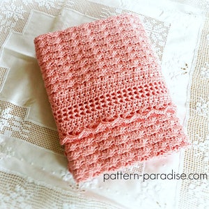 Puede incluir: Una manta de ganchillo doblada, de color coral, con un patrón texturizado. La manta presenta un borde festoneado y una sección calada decorativa. La manta descansa sobre una superficie blanca con adornos de encaje.