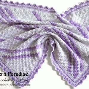 May include: A crocheted baby blanket in shades of white and purple with a scalloped edge. The blanket is folded in half and has a textured pattern. The text "Pattern Paradise Crochet by Maria pattern-paradise.com" is visible in the bottom right corner.