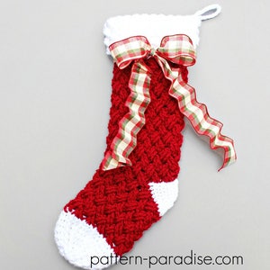 May include: A red and white crocheted Christmas stocking with a plaid ribbon bow. The stocking has a white cuff and a white toe. The stocking is hanging on a gray background. The text "pattern-paradise.com" is printed on the bottom of the stocking.