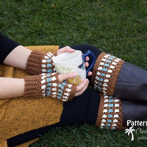 Könnte beinhalten: Eine Person trägt braune Lederstiefel mit gehäkelten Beinstulpen in Braun, Weiß und Blau. Die Person hält eine weiße Tasse mit einem blauen Henkel und einem Blumendesign. Die Person sitzt auf einer grünen Grasfläche.
