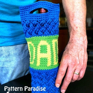 May include: A blue and green crocheted wine bottle holder with the word "DAD" in green and yellow yarn. The holder is being held by a person's hand.
