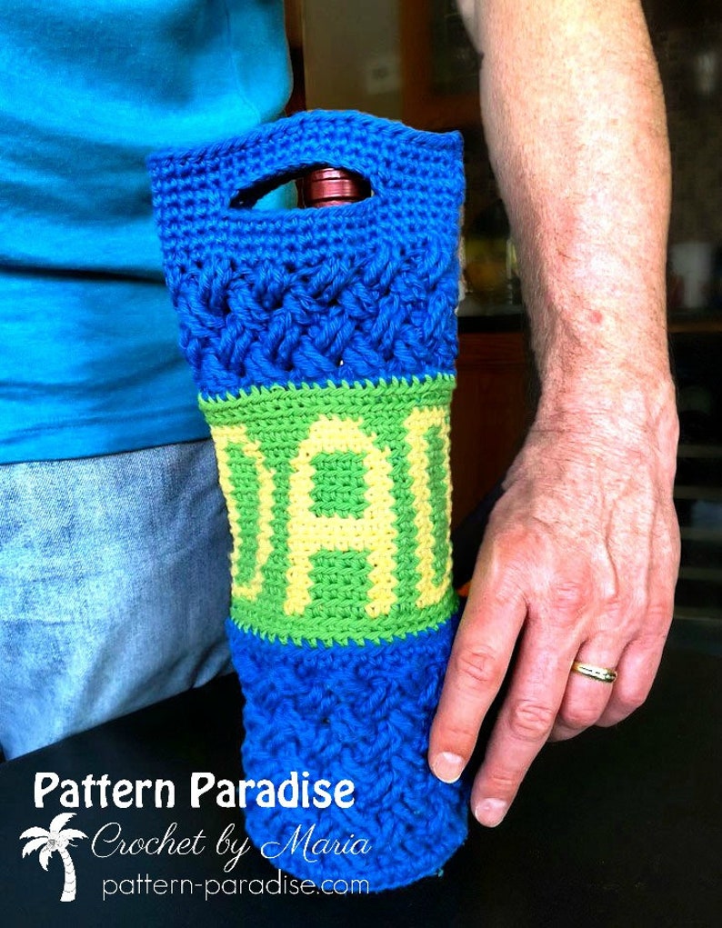 May include: A blue and green crocheted wine bottle holder with the word "DAD" in green and yellow yarn. The holder is being held by a person's hand.