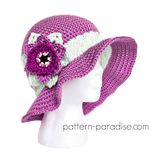 May include: A handmade crochet sun hat in shades of purple and white, adorned with a large purple flower. The hat features a wide brim and is displayed on a white mannequin head. The text "pattern-paradise.com" is visible.