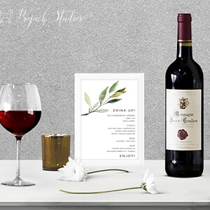 Puede incluir: Una tarjeta blanca con una ilustración de una rama verde frondosa y el texto "Drink Up!" con una lista de bebidas recomendadas, incluyendo licores, vino y cerveza. Una botella de vino tinto y una copa de vino tinto están sobre una superficie blanca.