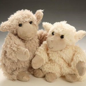 Könnte beinhalten: Zwei Plüsch-Lamm-Spielzeuge, eines beige und eines weiß, sitzen nebeneinander. Sie haben flauschiges Fell und schwarze Augen.